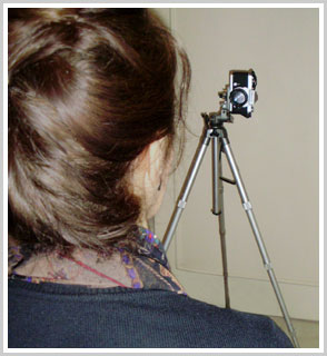 Modelo feminino, cabelo preso, sentada de costas, na sua frente está a câmera sobre tripé
